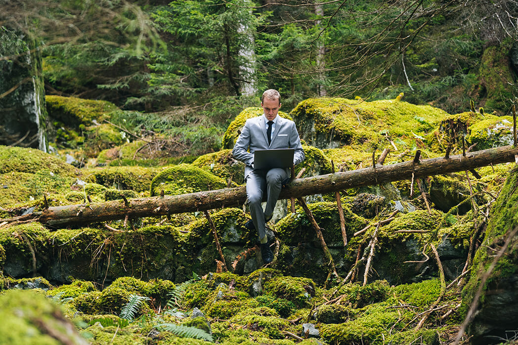 Ein Mann im Anzug sitzt auf einem Baum im Wald mit einem Laptop auf seinem Schoß.